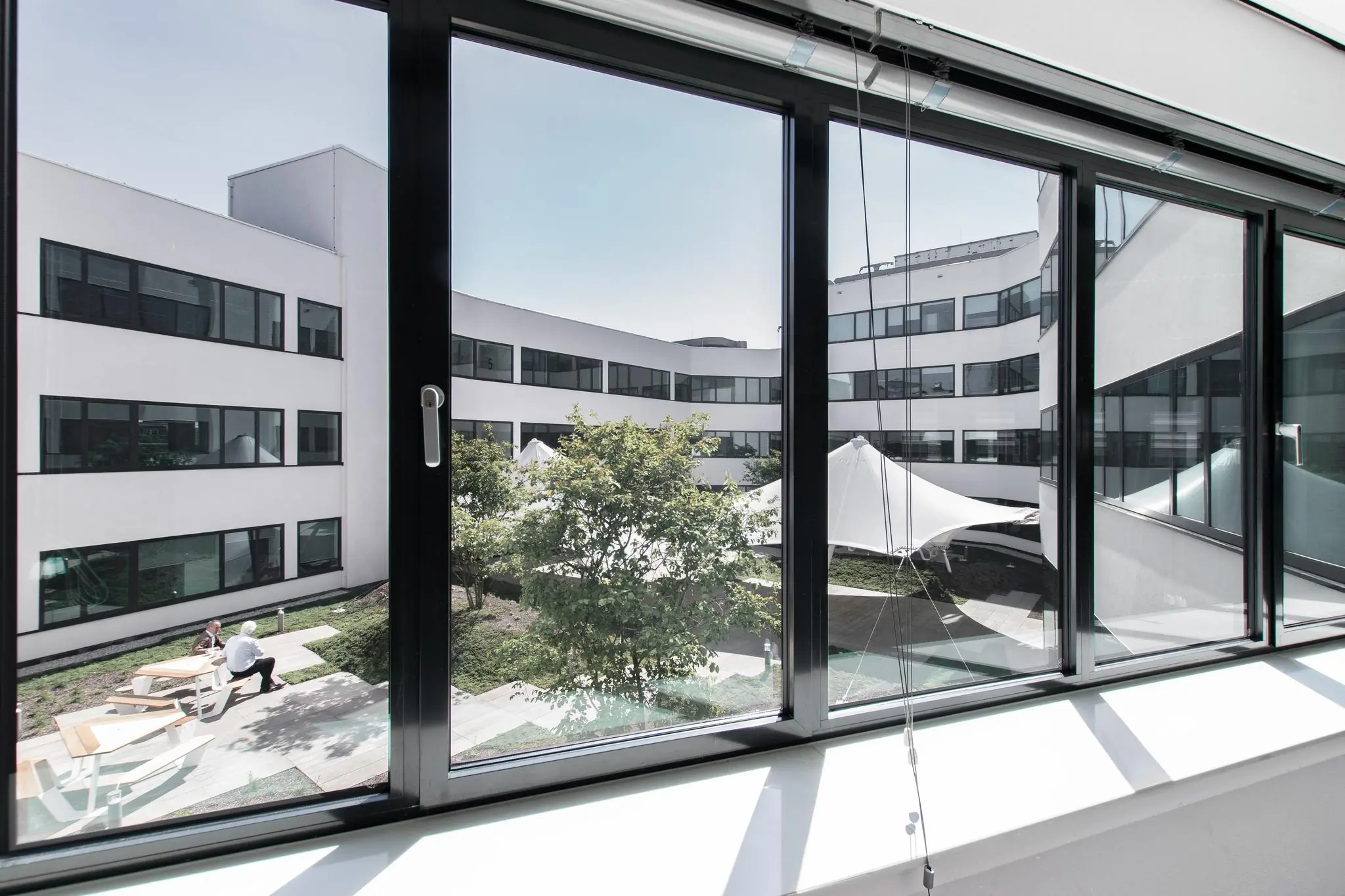 Uitzicht door een raam op een moderne binnenplaats met witte gebouwen, bomen en zitplekken in de Grote Marktstraat.